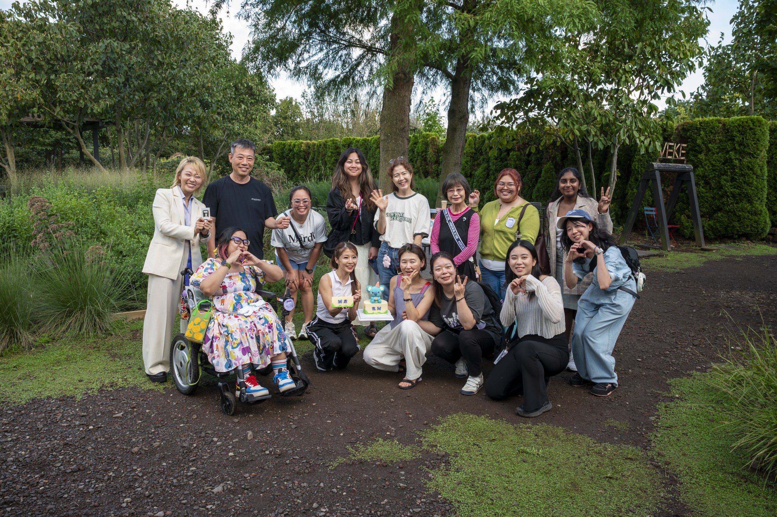 Group photo with two rows totalling a dozen people. The back row stands; front row mostly squats, and includes one wheelchair user. Most people wave/make peace or other hand signs. Two hold the blue koala cake. Background a lush green garden.
