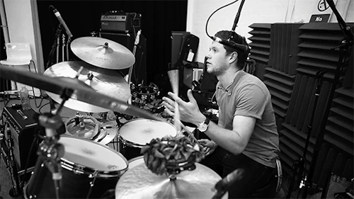  An animated gif of Niall Horan sitting at a drum set and spinning a drumstick between his fingers. 