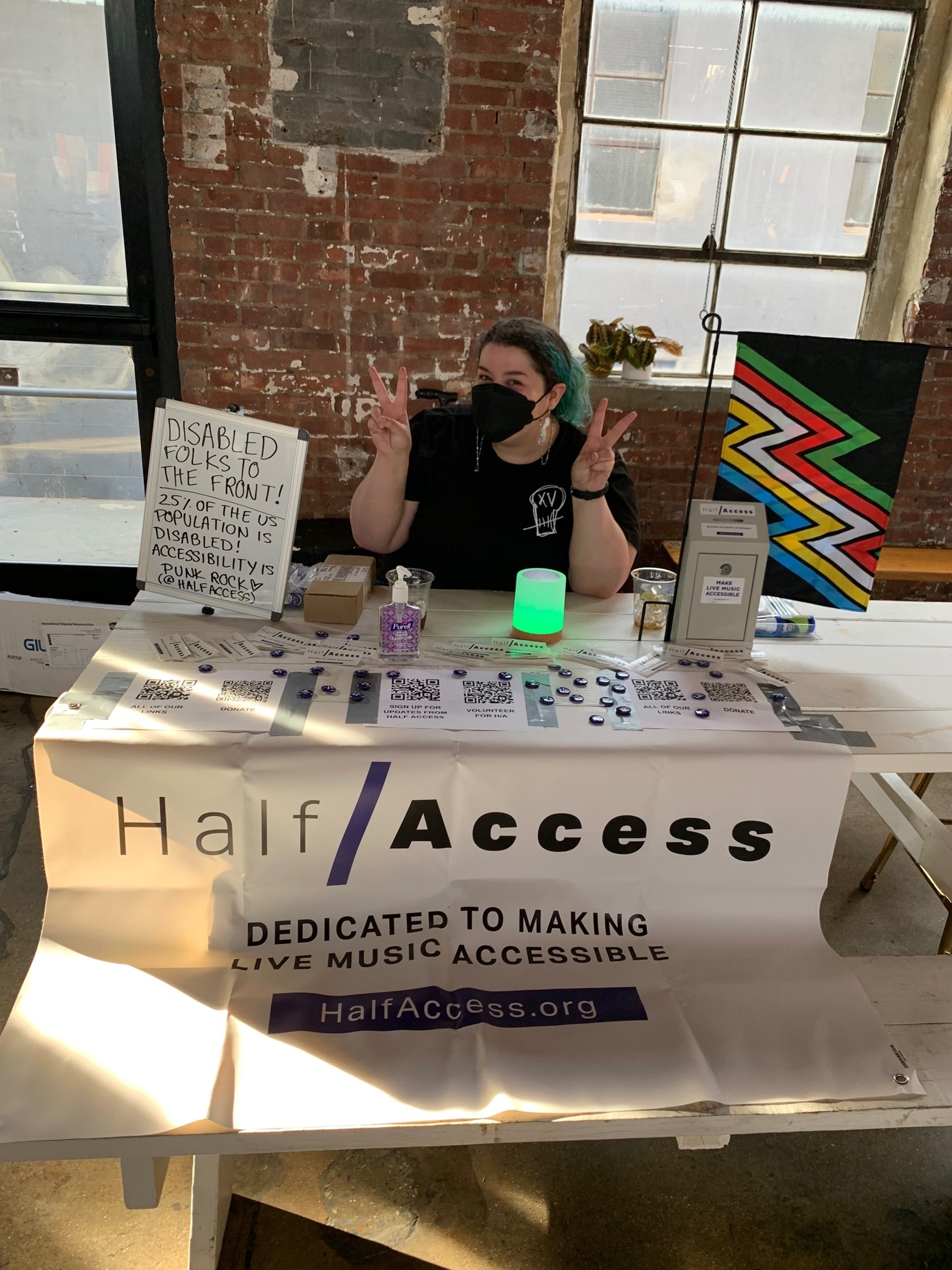 Photo of Valerie sitting at a table making peace signs with her hands. Banner on table reads: Half/Access Dedicated to Making Live Music Accessible HalfAccess.org.