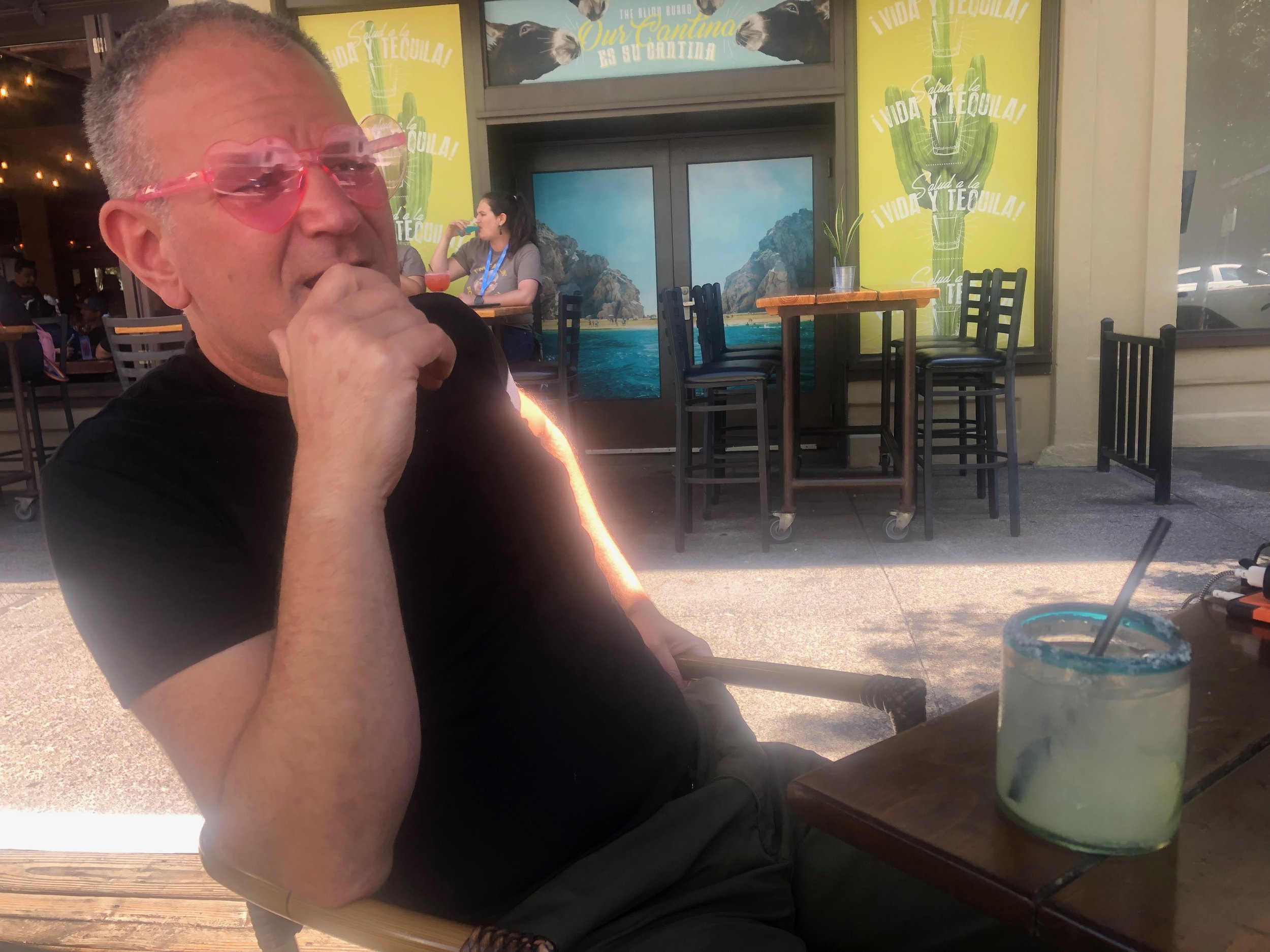 Photo of Javi in heart-shaped Barbie glasses sitting at an outdoor table, hand on his face