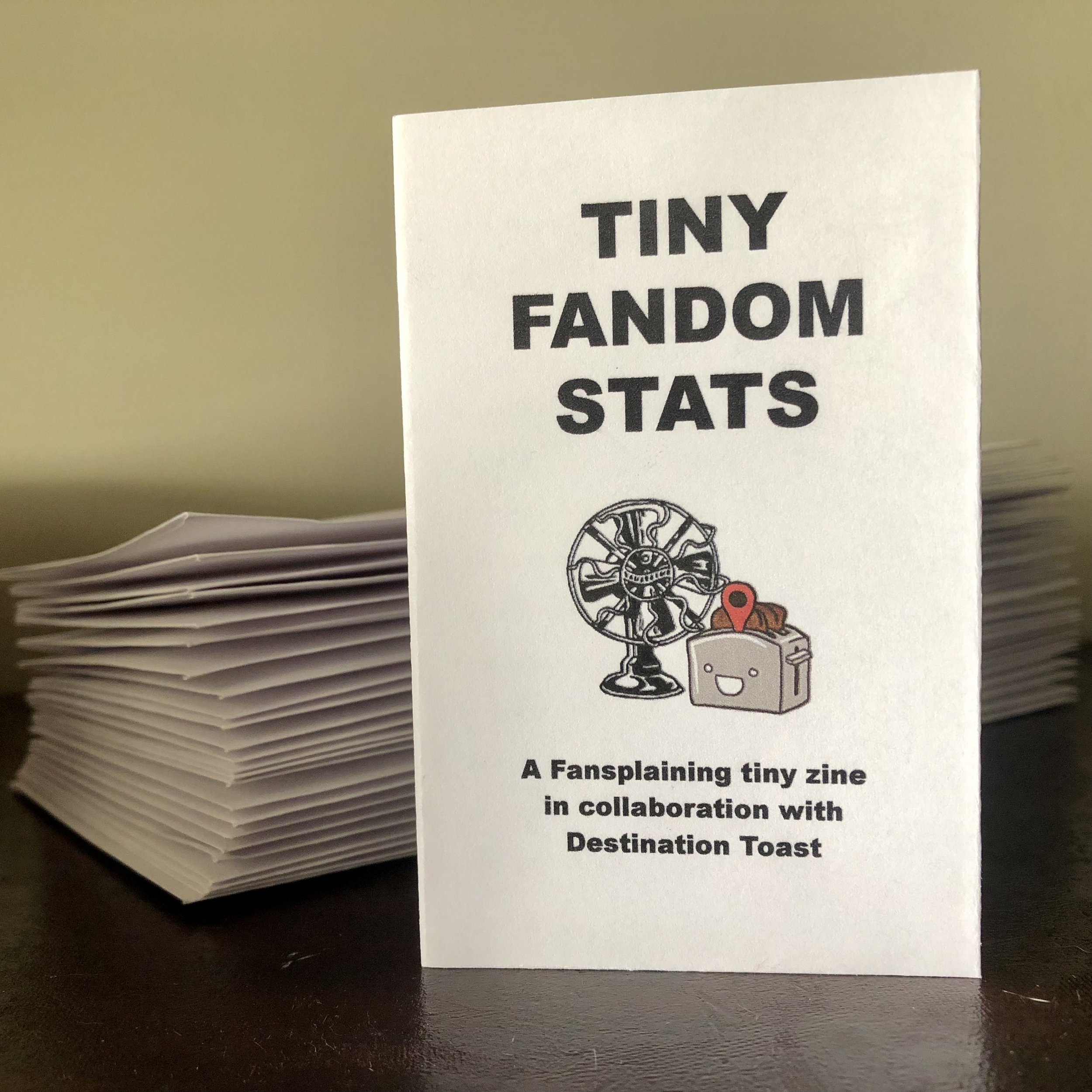 A stack of tiny zines and one upright. The title reads "TINY FANDOM STATS" followed by an image of the Fansplaining fan and Toast's toaster/toast avatar. The subtitle below reads "A Fansplaining tiny zine in collaboration with Destination Toast."