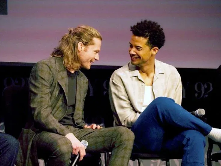 Photograph of Sam Reid and Jacob Anderson sitting onstage holding mics, smiling at each other.