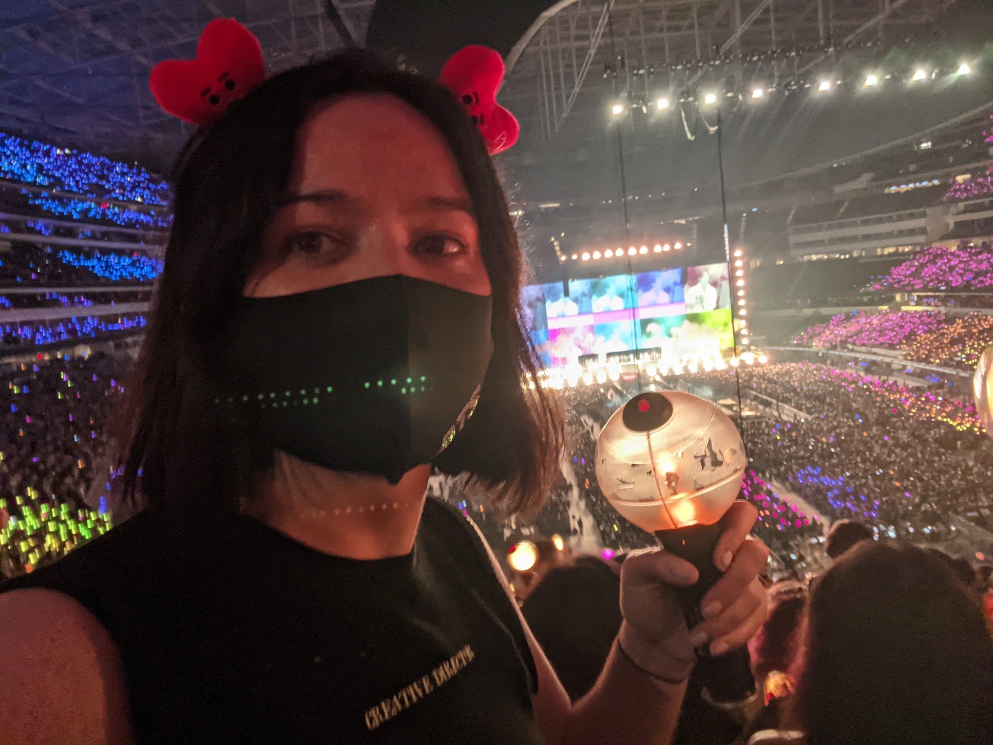 Photograph of Kayti at a Kpop concert, wearing a mask and a headband with little hearts as ears, and holding a lightstick.