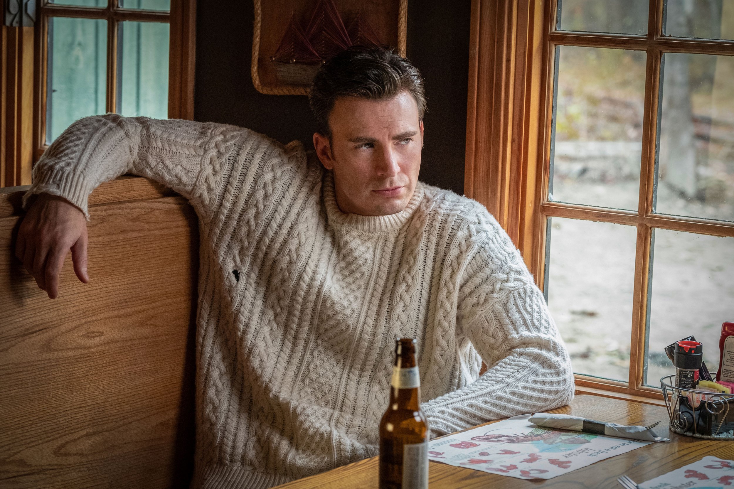 Photo of Chris Evans as Ransom Drysdale from Knives out in That Sweater, sitting in a wooden booth at a restaurant, bottle of beer in front of him