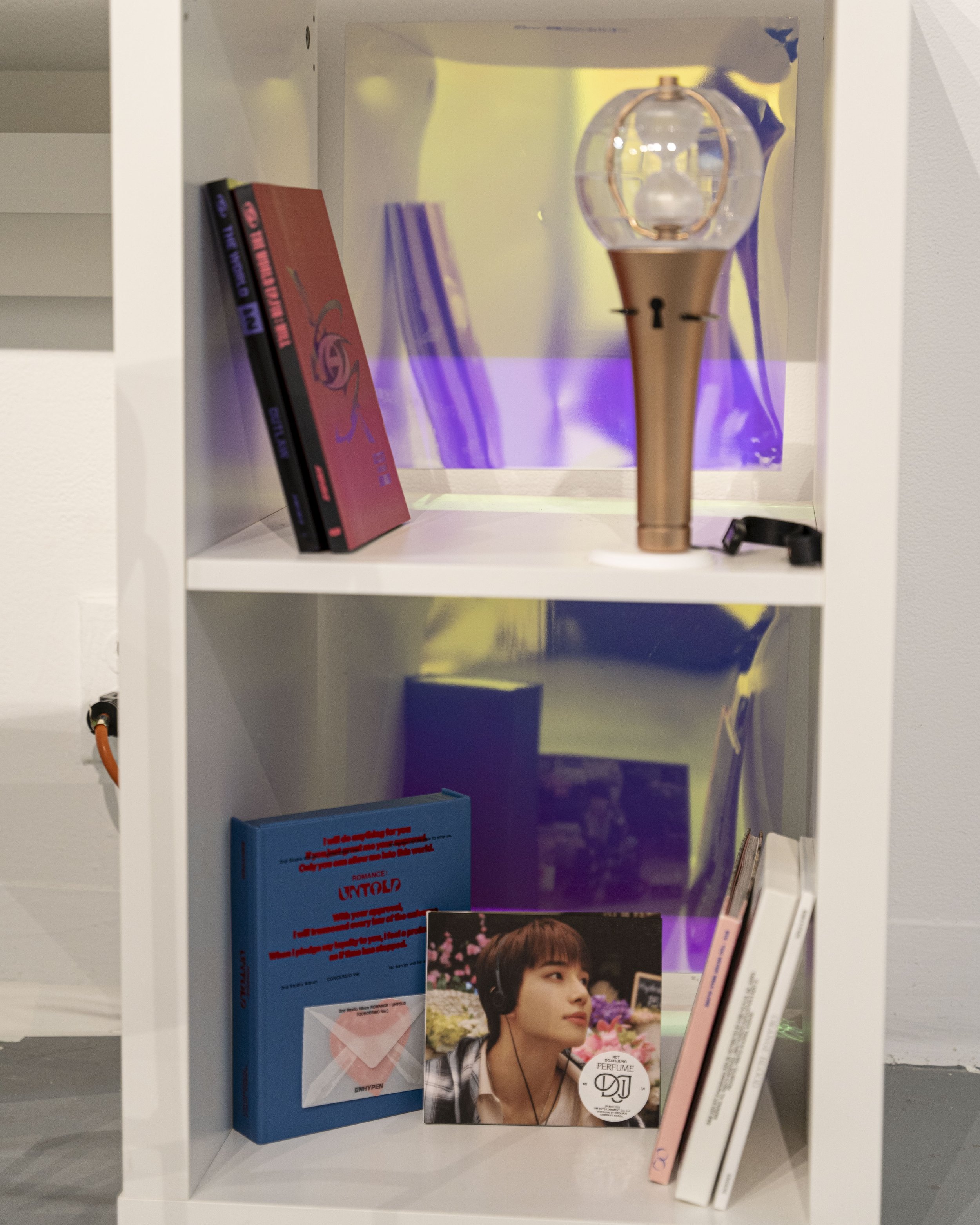 A collection of K-pop items on white IKEA shelves, with a light stick on the top shelf and several albums on the bottom.