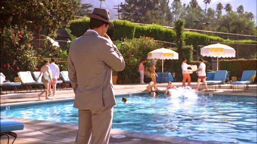 Still from Mad Men featuring Don in a light-colored suit and hat with his back to the camera, looking at people in and around a pool
