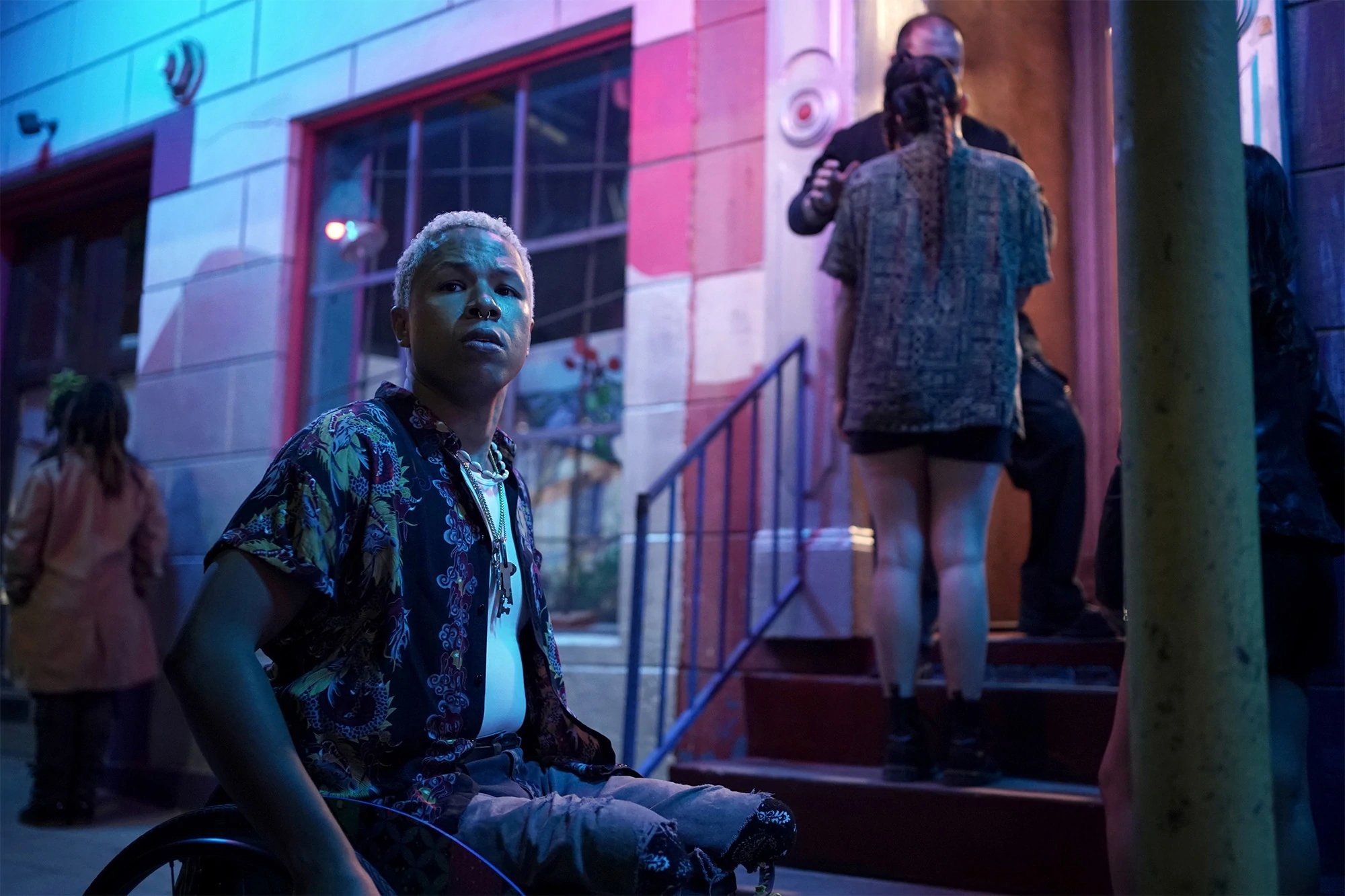 Photograph of Marvin from Queer as Folk in his wheelchair on a sidewalk in front of a building; two figures stand on the steps of the building