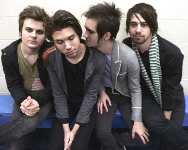 Photograph of the four members of Panic! at the Disco sitting very close to each other on a blue bench.