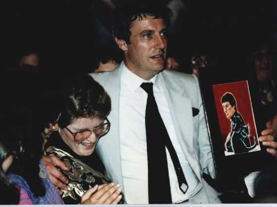 Photograph of Paul Darrow in a light-colored suit with his arm around Leah Rosenthal, who's smiling and wearing glasses. A piece of fanart is in Paul's hand.