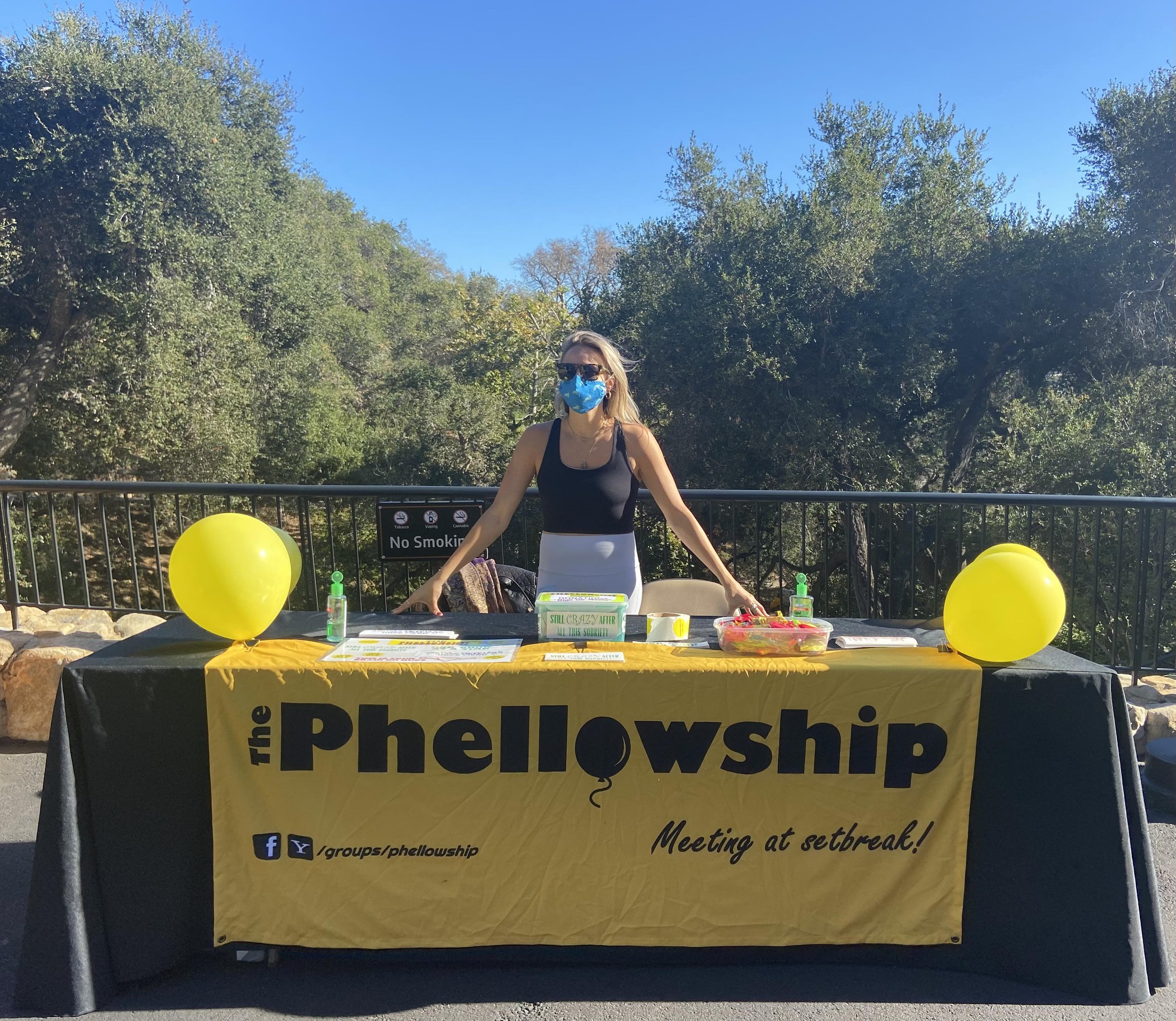 Photograph of a woman in a mask at an outdoor table with a big yellow Phellowship sign and several yellow balloons