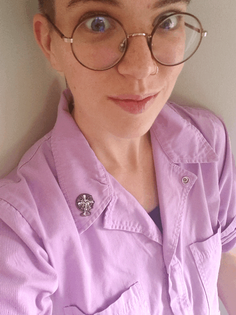 Animated gif of Flourish pointing to the fan-shaped enamel pin on their jumpsuit lapel