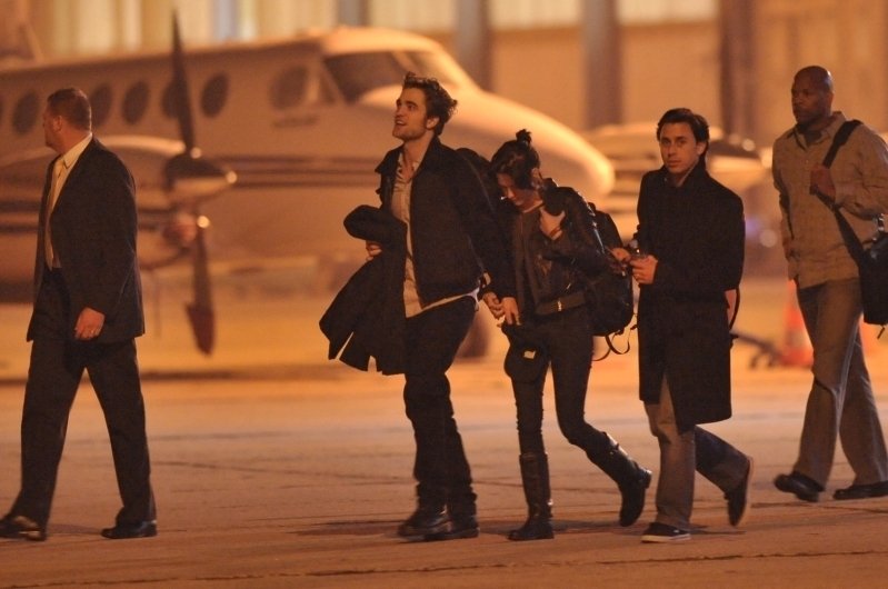 Photograph of Robert Pattinson and Kristen Stewart photographed at an airport in France holding hands. Several figures flank them, and a plane is prominent in the background.