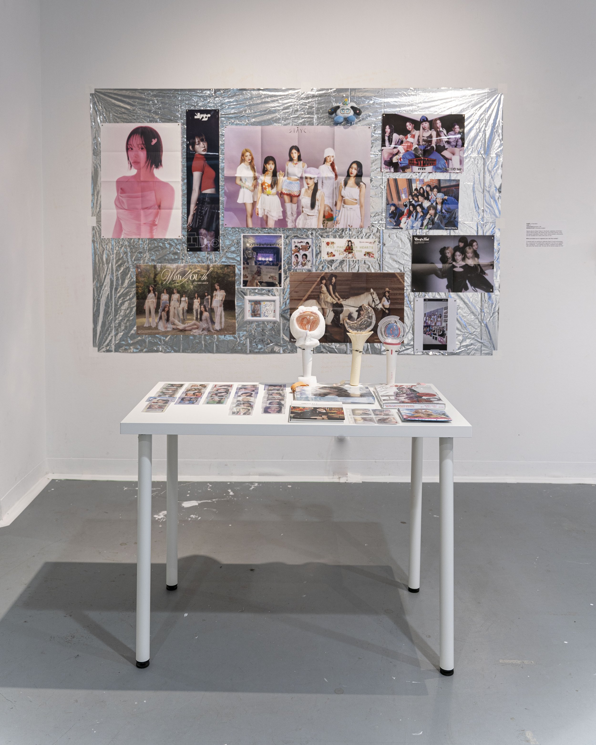 Photograph of a collection of K-pop fandom items including photo cards, light sticks, and, in a collage on the wall against a silver background, a selection of posters.