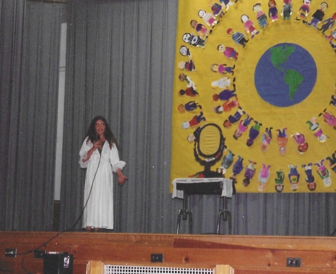 Elizabeth, in the fifth grade, wearing a white nightgown and singing into a mic with an incredibly 1990s backdrop.