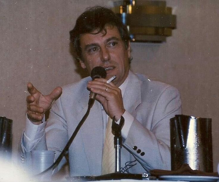 Photograph of Darrow in a light colored suit and tie, seated at a table, holding a mic and a cigarette in the same hand.