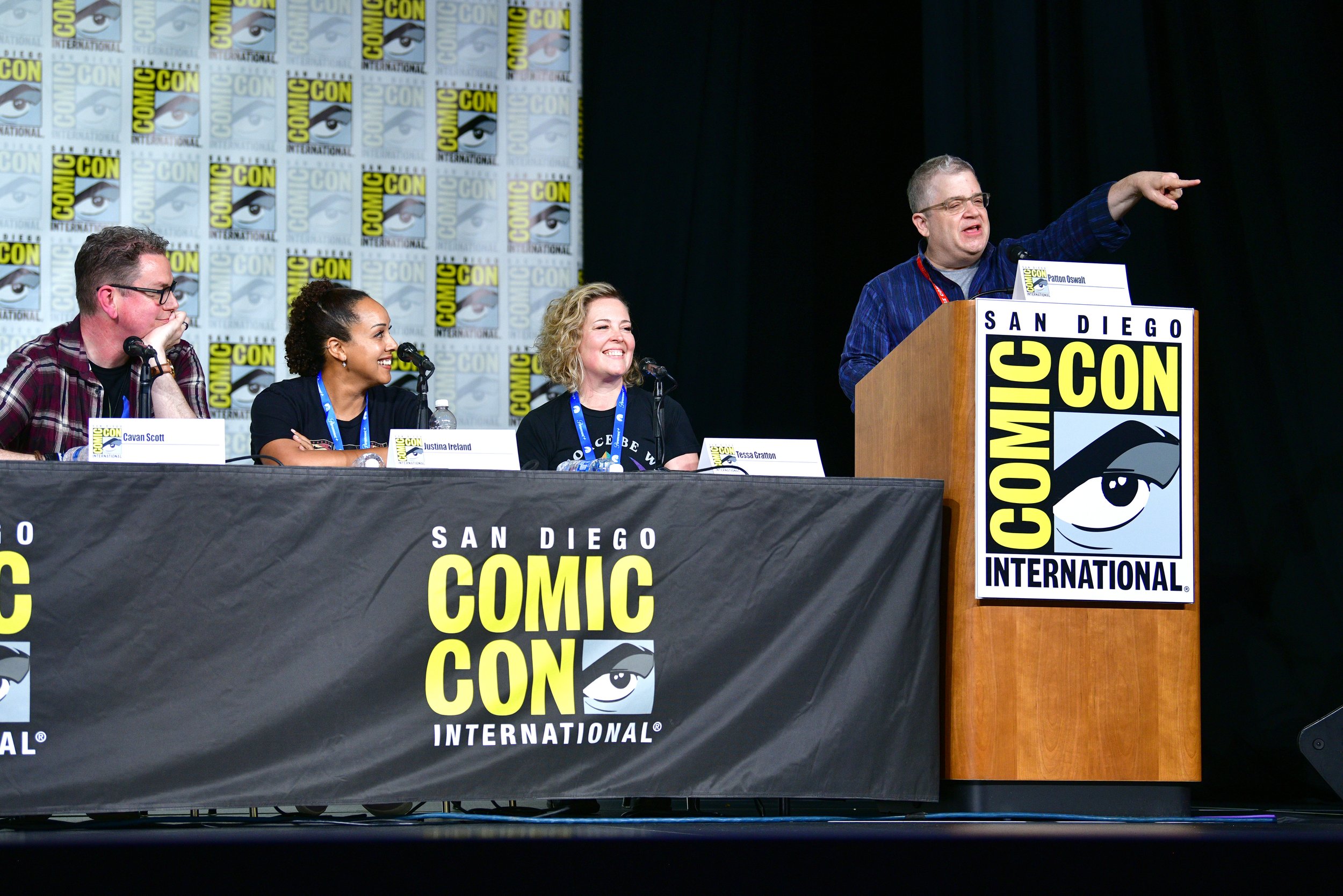 Photograph of a panel on the stage at SDCC: Cavan, Justina, and Tessa seated, Patton standing and pointing at the dais.