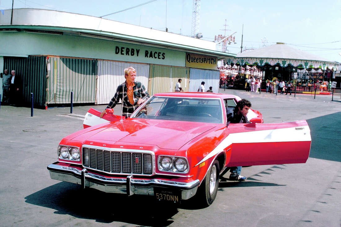 Still from the show depicting Starsky and Hutch getting out of their red and white Ford Gran Tarino, a carnival scene (derby races and a merry-go-round) in the background.