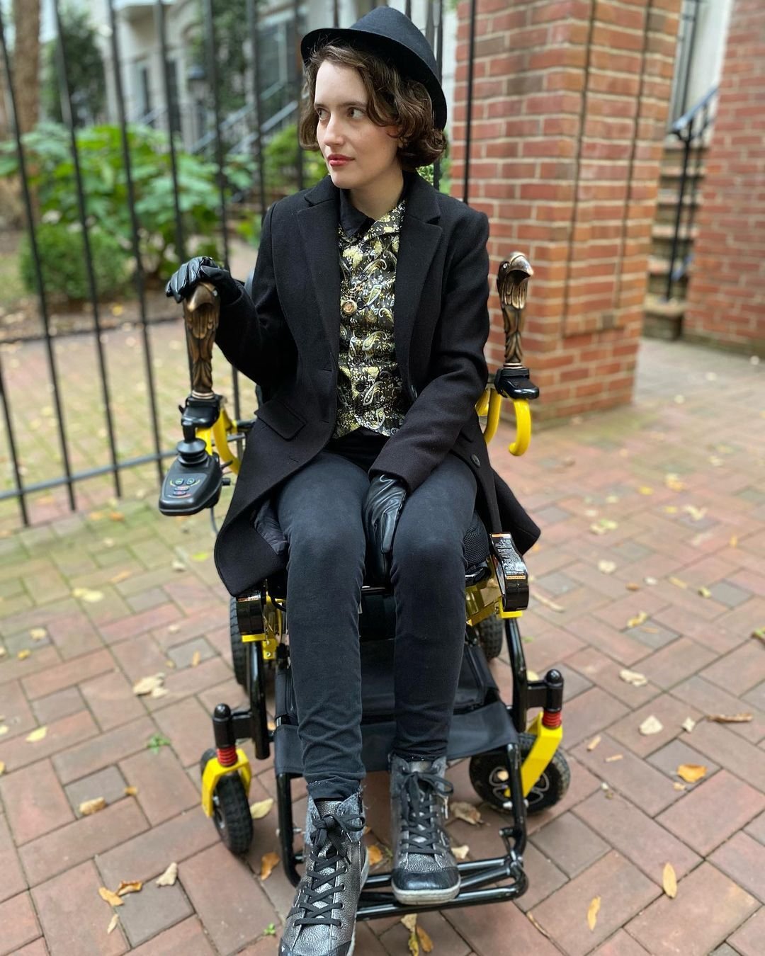 Figure in a hat, long coat, waistcoat, and leather gloves, seated in a wheelchair with bronze bird heads on its armwrests