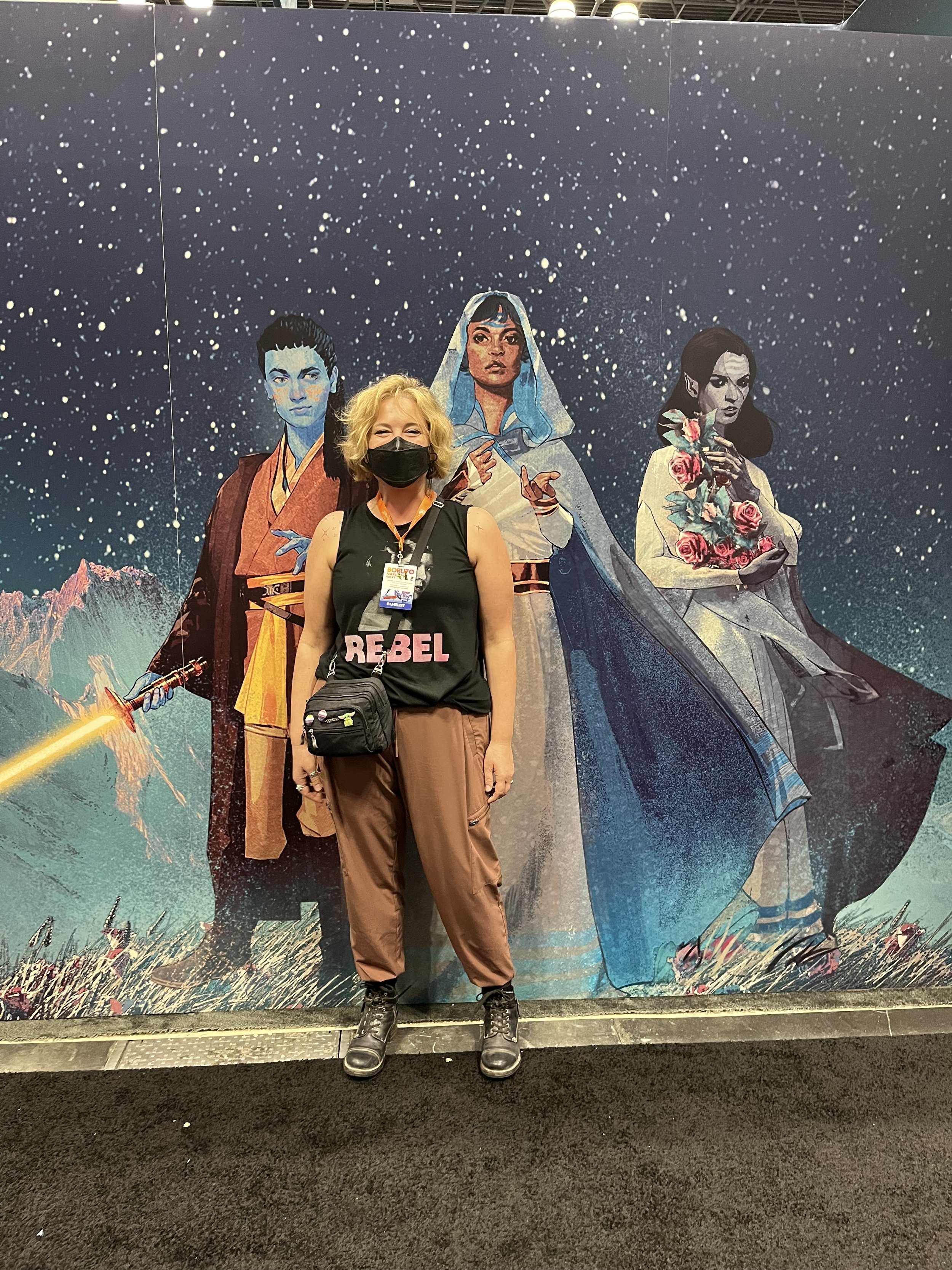 Photograph of Tessa standing in front of a full-wall-sized version of cover art from their book, featuring three Star Wars characters.