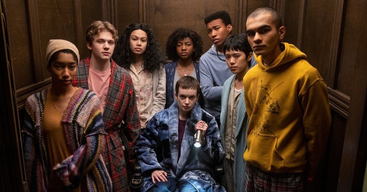 Still photograph from Midnight Club featuring seven young people in pajamas and robes looking directly at the camera. The figure in the center is seated in a wheelchair and holding a flashlight.