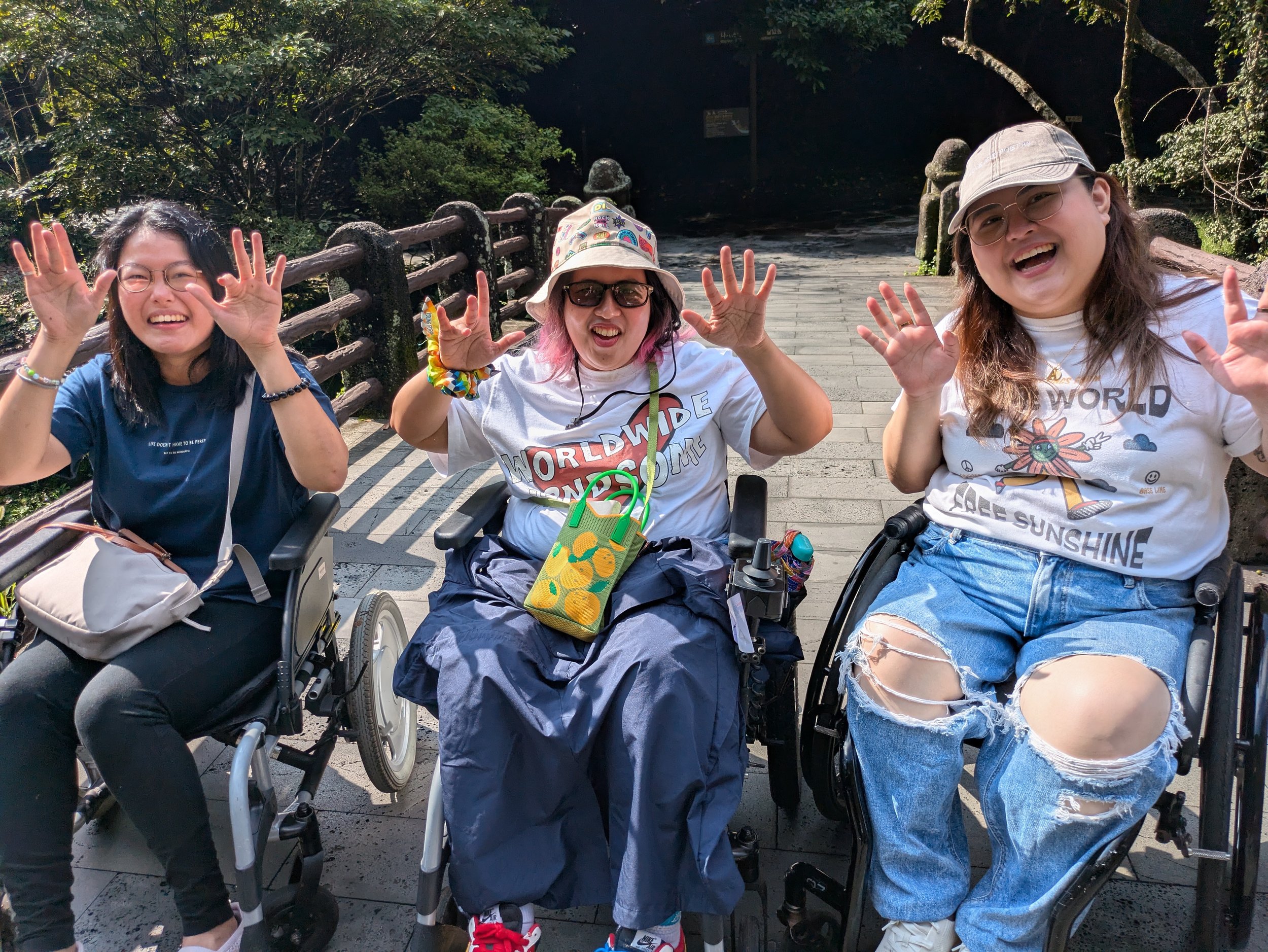 Yu Qi,From left to right, Yu Qi, Karlina, and Aia, all sitting in wheelchairs and waving to the camera, situated on a stone bridge.