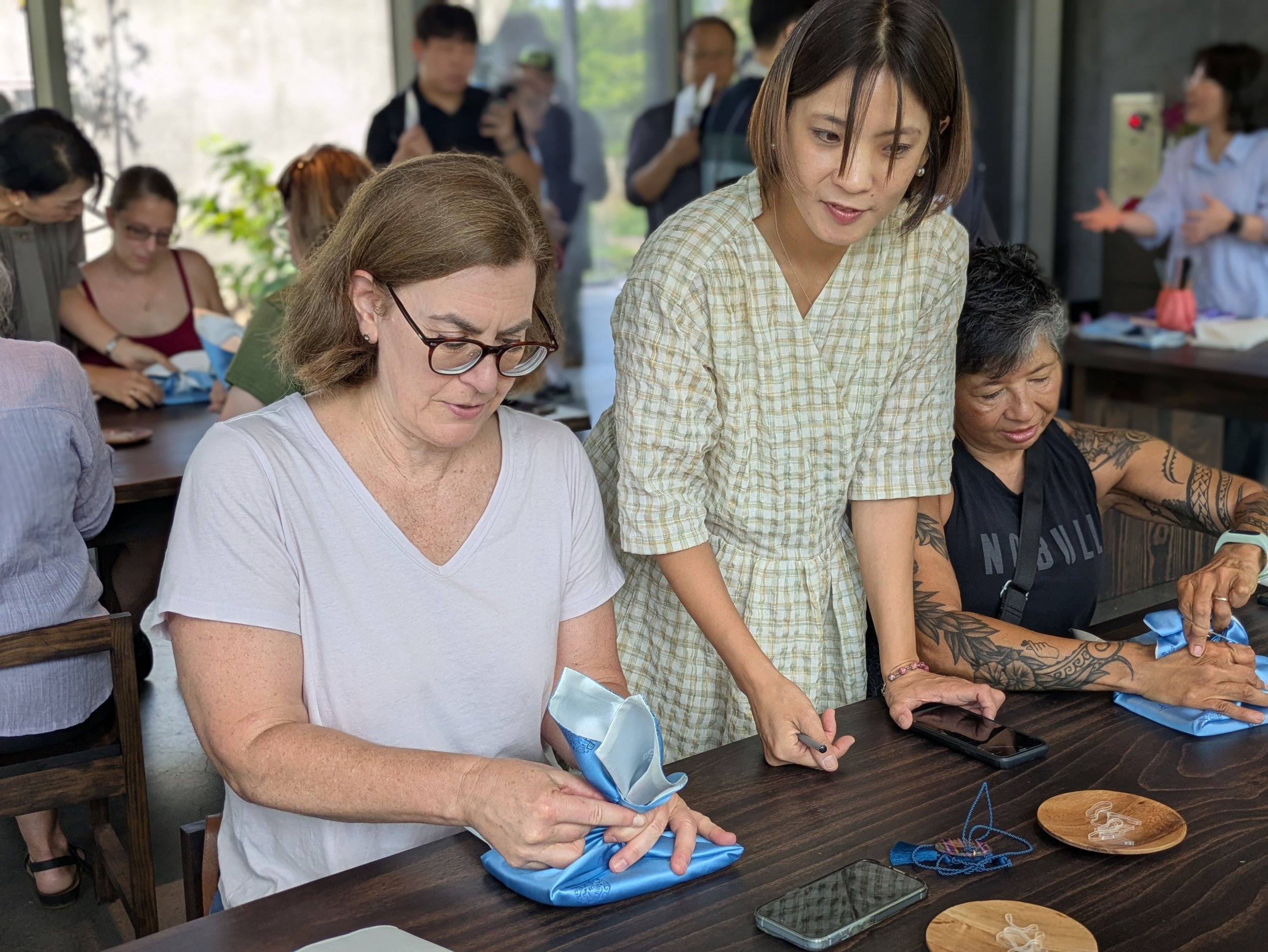 Image of two Purple Festa attendees sitting at a table wrapping books in blue cloth, with an instructor standing between them.