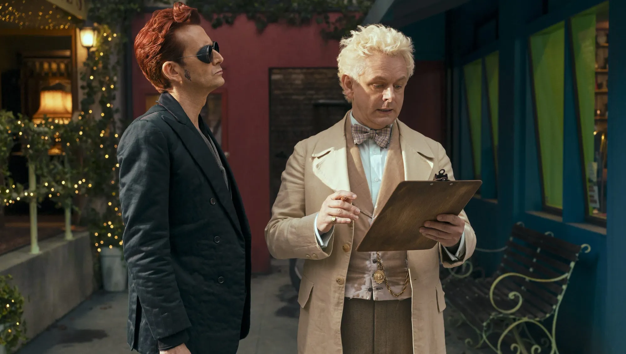Crowley stands looking at Aziraphale, who is looking at a clipboard.
