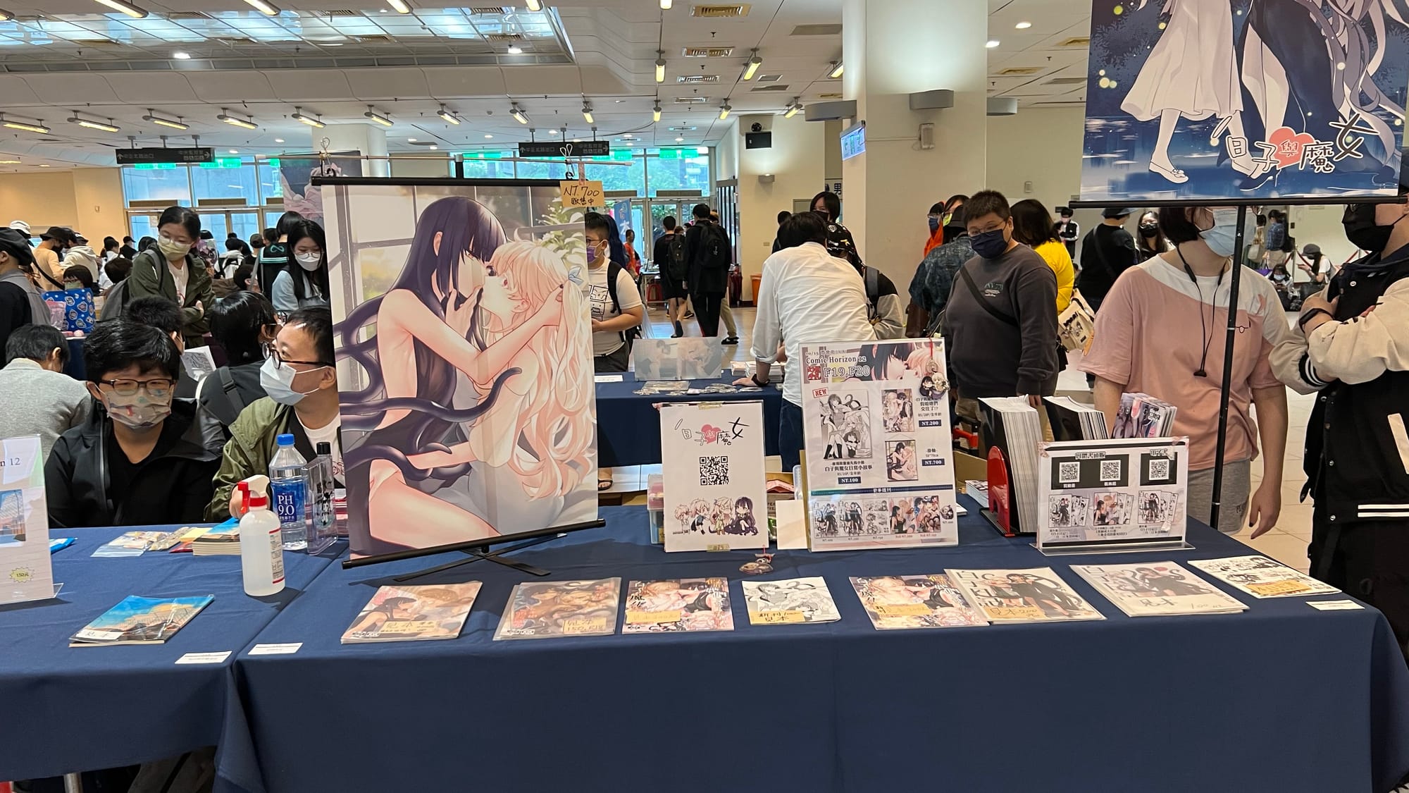 A table on a showroom floor featuring racy comics (doujinshi) spread out in a row, with flyers behind as well as an illustrated erotic poster of two scantily clad girls of indeterminate age kissing.