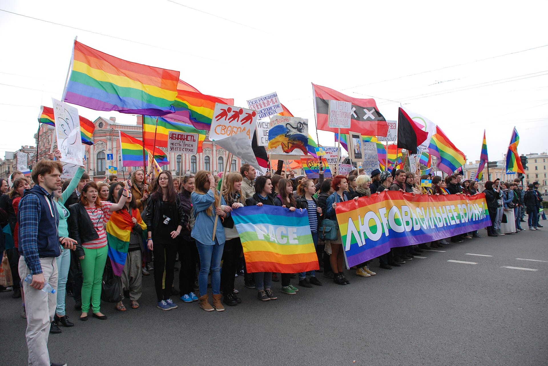 A large crowd of protesters standing on a city street holding pride flags and other flags, some of them chanting.