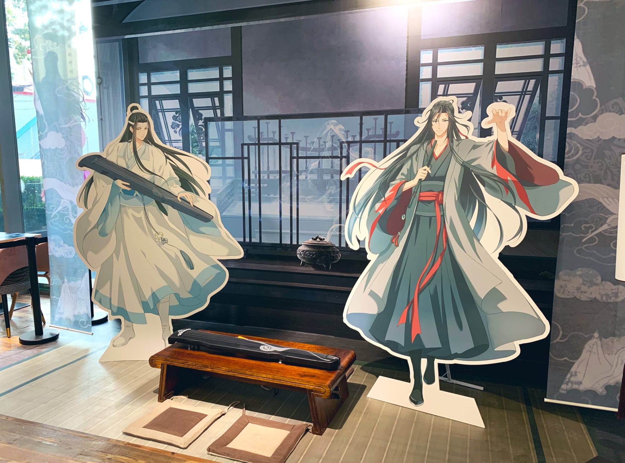 A display of two famous characters from Mo Dao Zu Shi, featuring standees of each illustrated character in their flowing robes, a raised dais featuring a guqin, and a lit incense burner sitting before a decorative background.