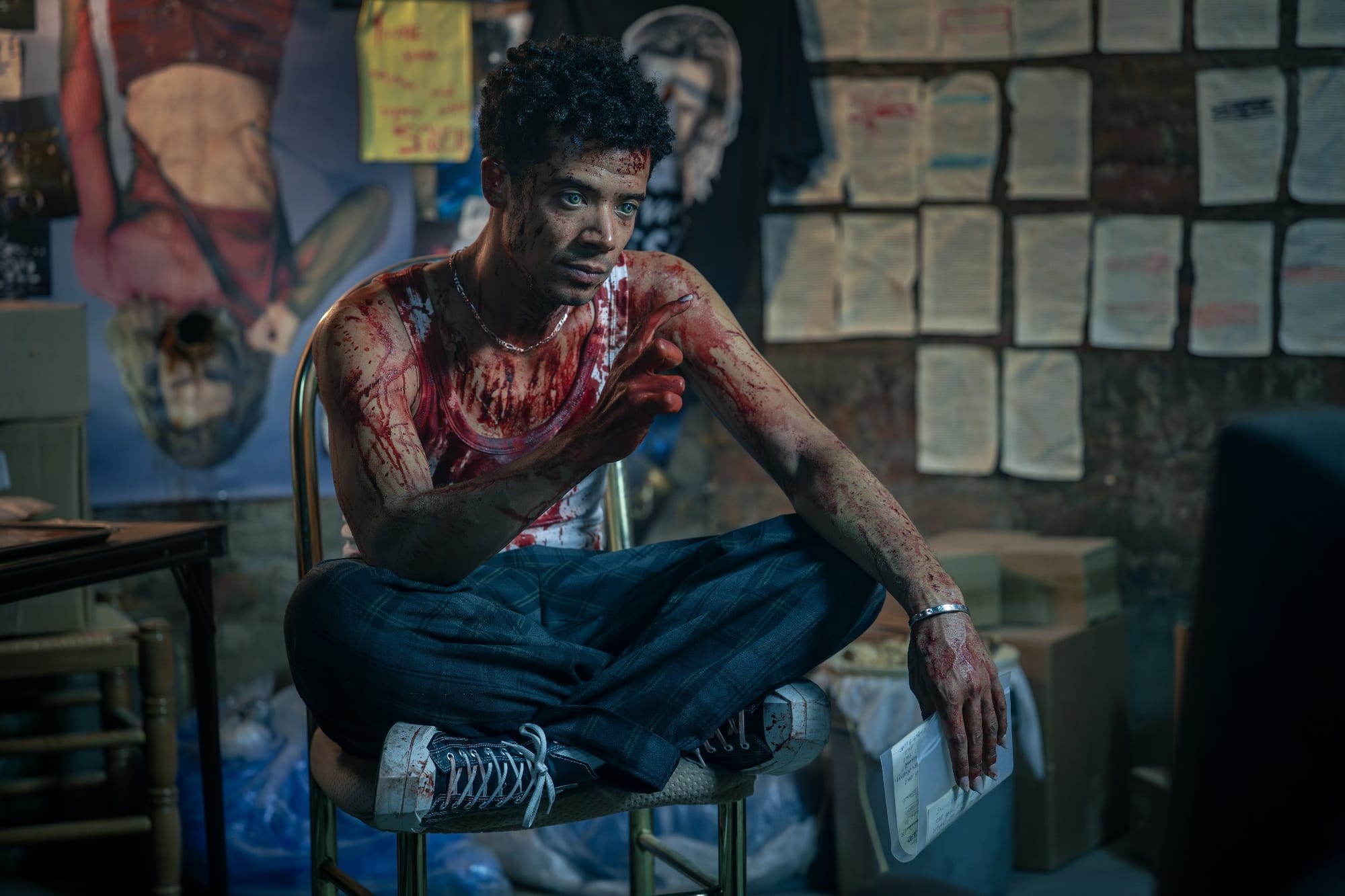 Production still featuring Louis, covered in blood, pointing his finger as he sits cross-legged on a chair.