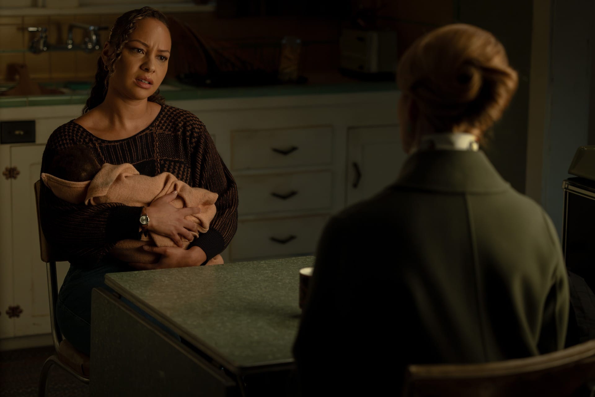 Production still of Bernadette with Baby John in her arms, sitting at a kitchen table across from Guardian Laura Linney, who has her back to the camera.