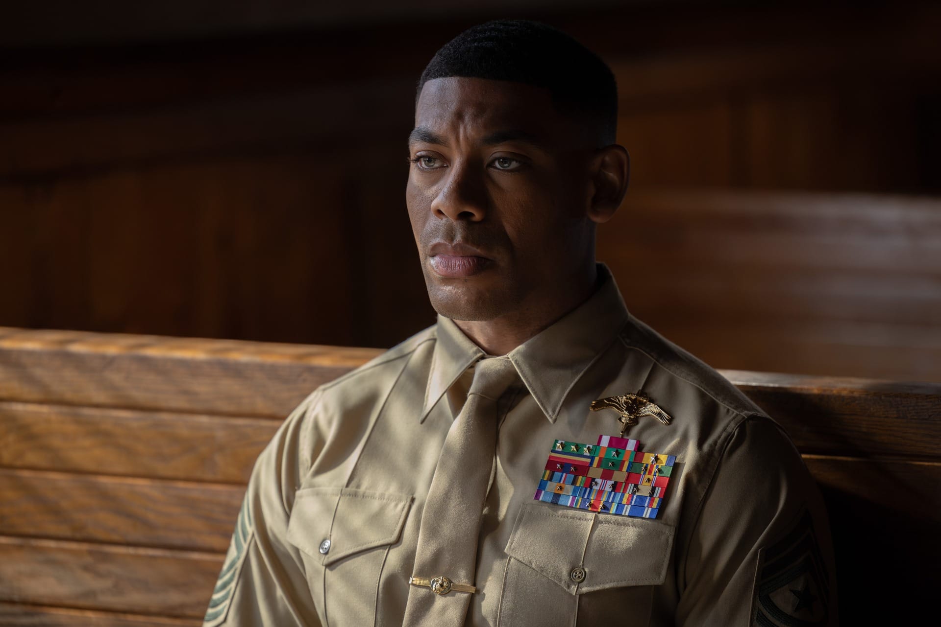 Production still of John in a tan Marines uniform, looking stone-faced as he sits on a wooden bench.