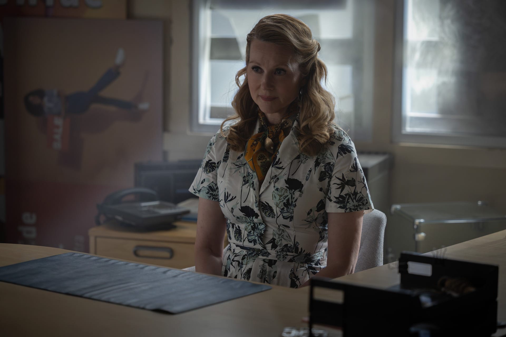 Production still of Guardian Laura Linney in a white floral dress, sitting behind a desk.