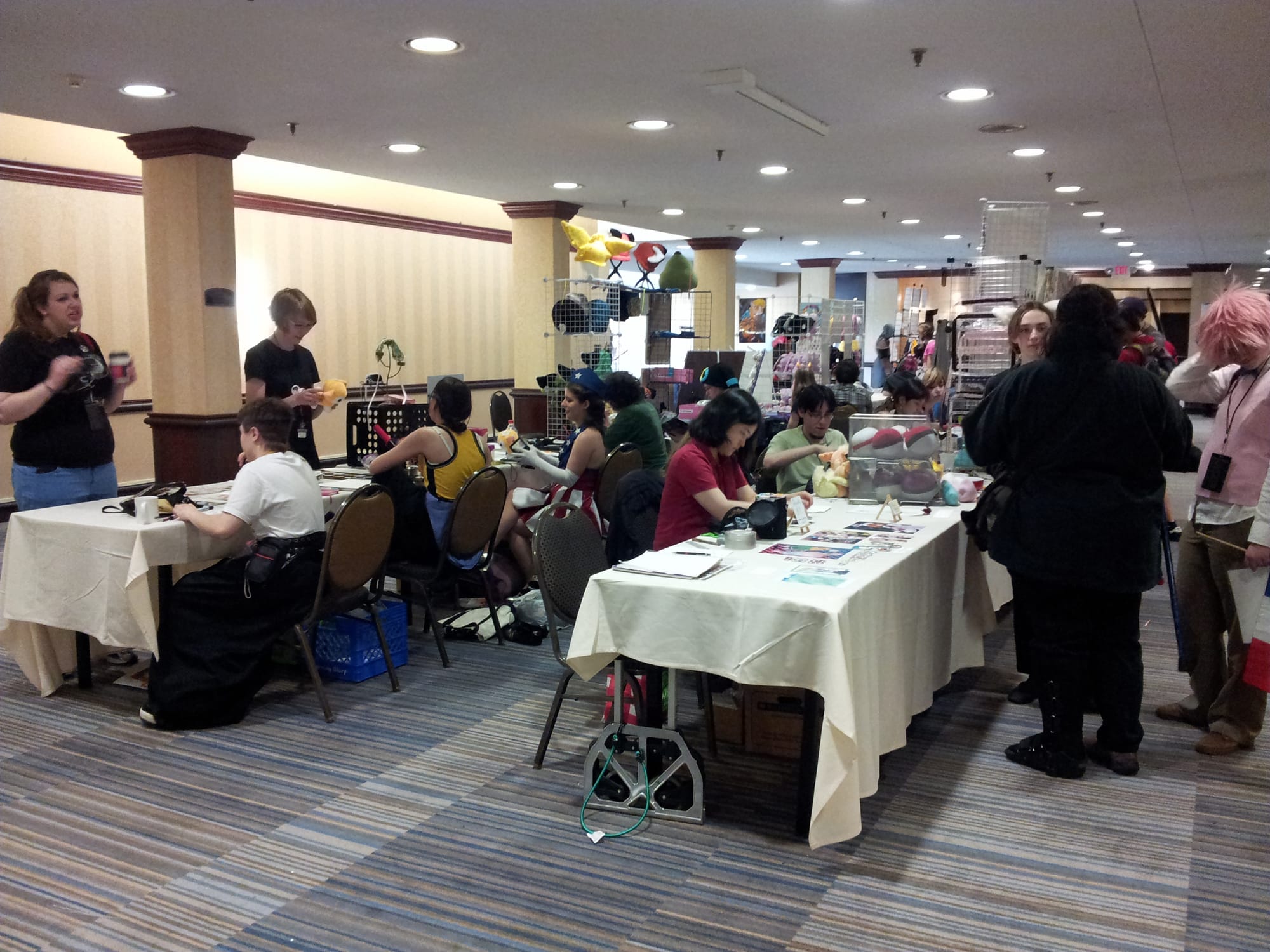 A hotel basement is turned into an artist's alley with tables and anime fans. It's not crowded, and many artists are talking to the customers in front of their table. There are very few "booth" setups.