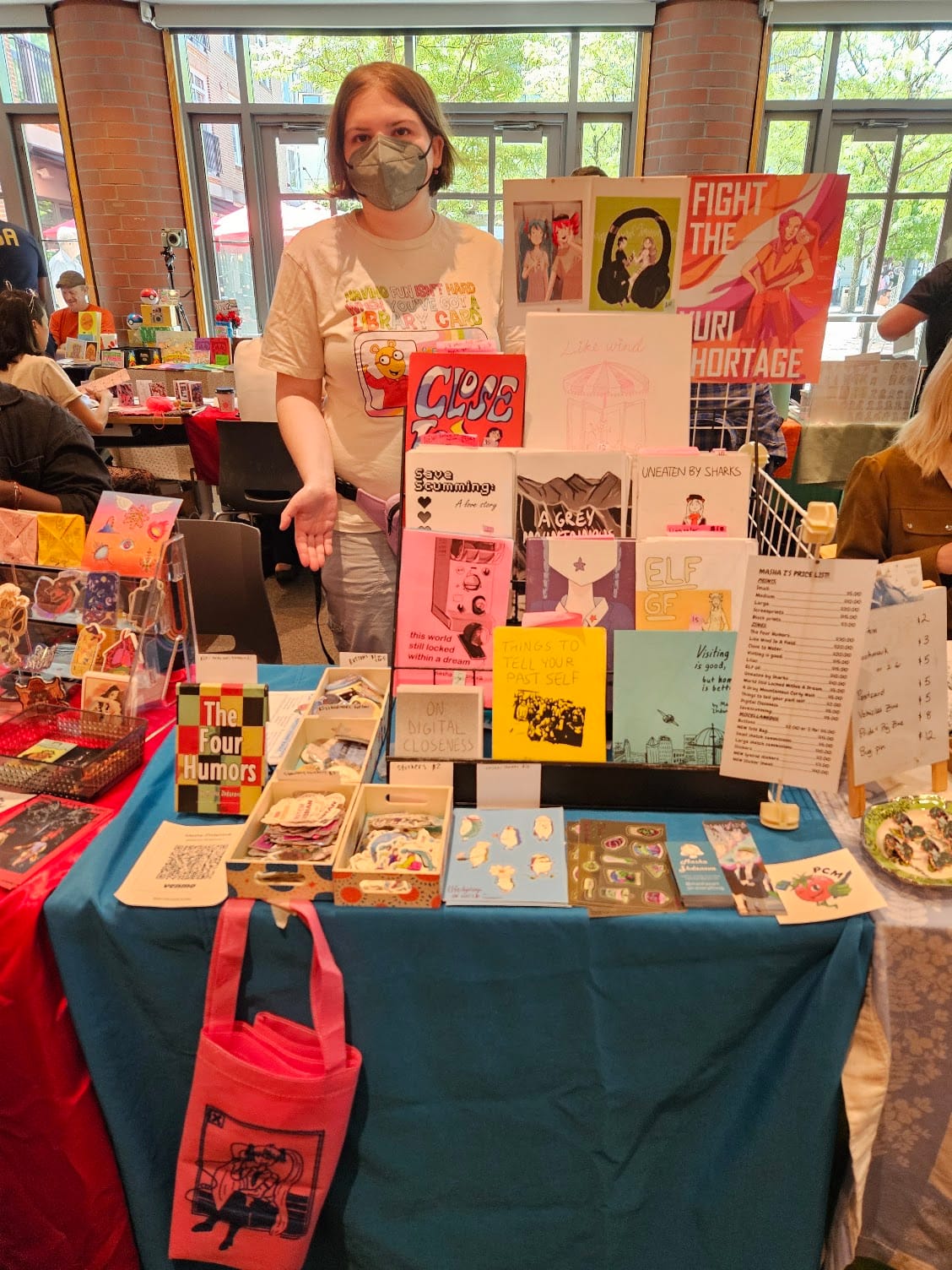 A young artist in a white Arthur shirt and grey mask shows off her table at a Zine fest. The table is small, but full of stickers, zines, and a couple posters.