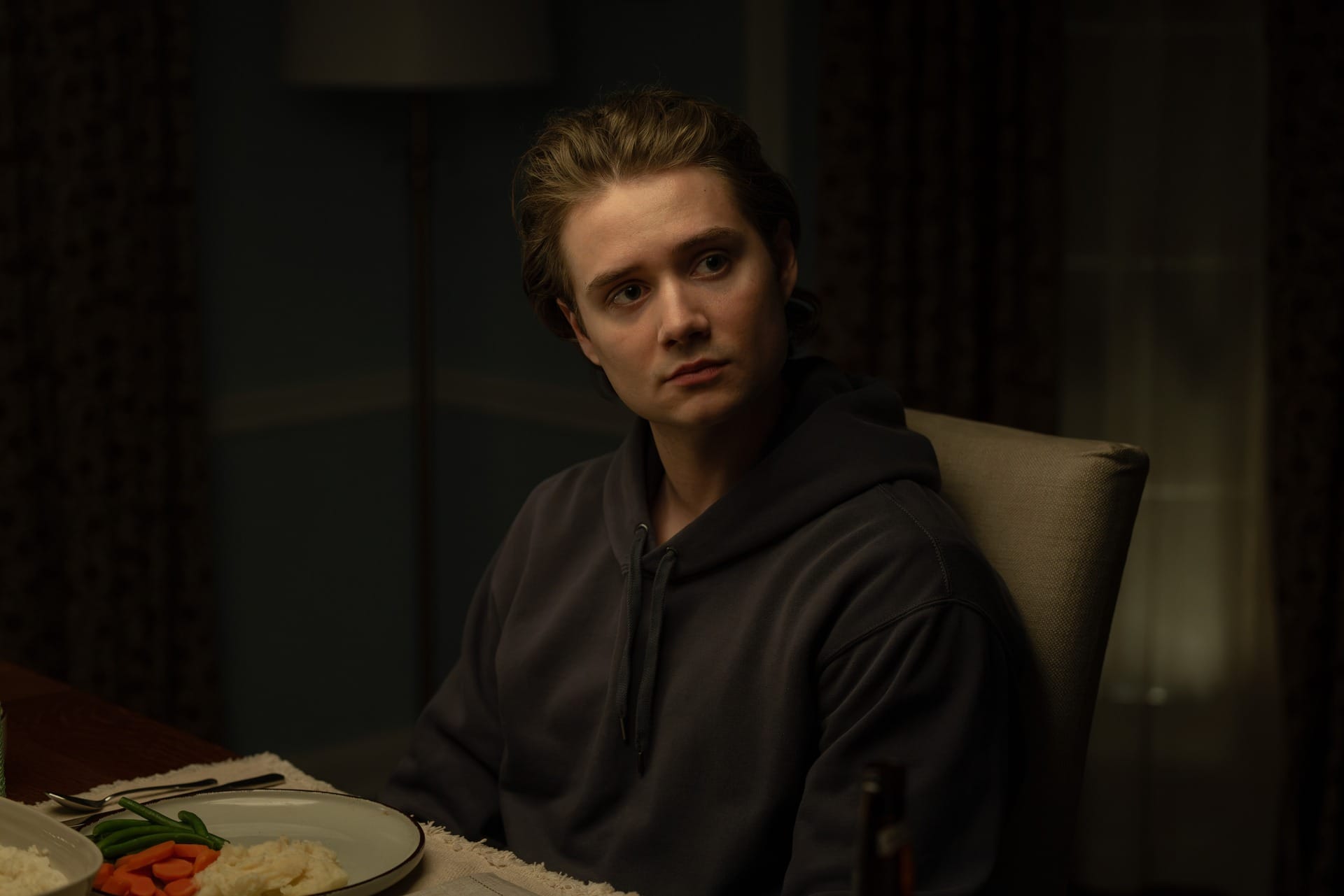 Production still of Noah sitting at a dining table in front of a plate of vegetables.