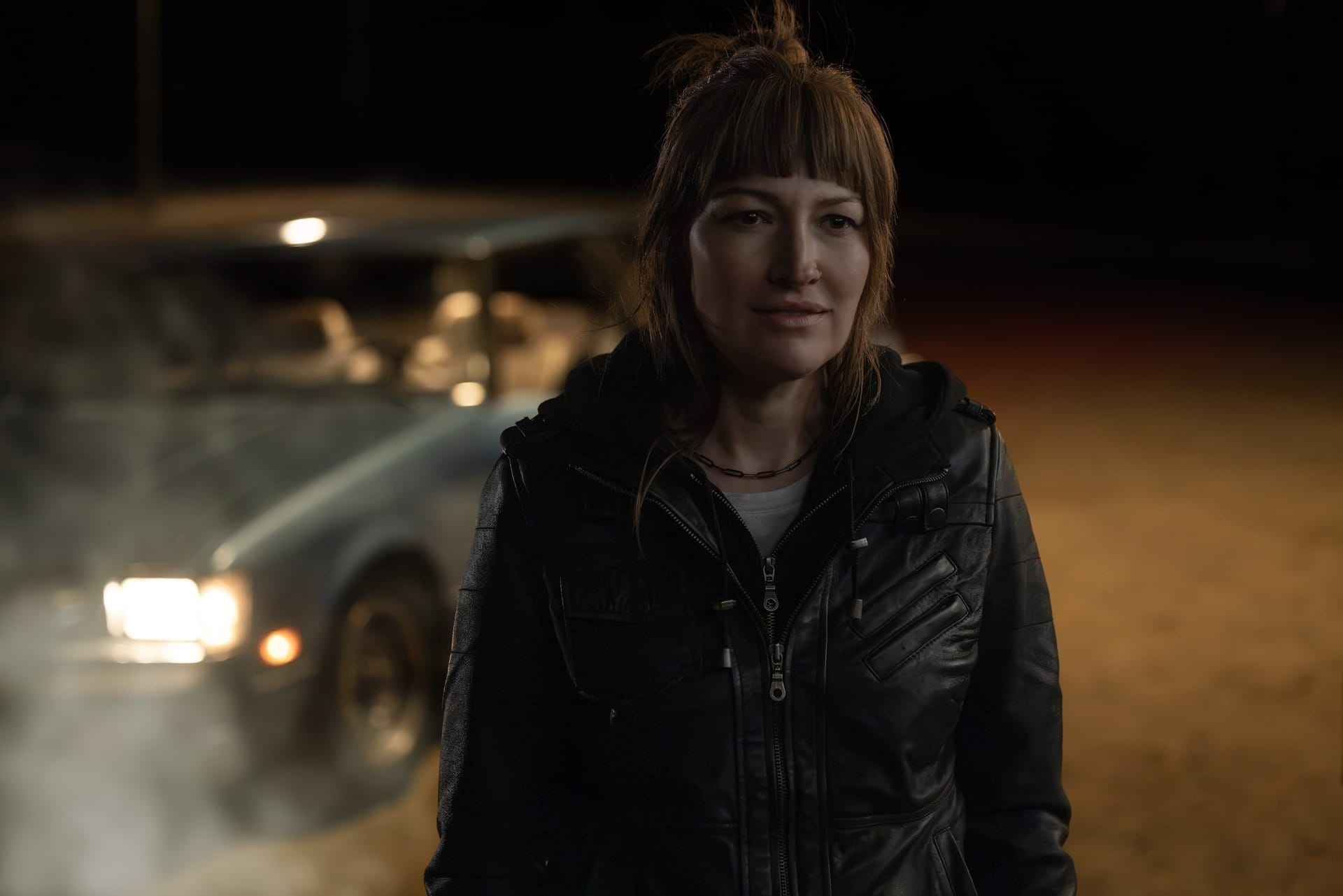 Production still of Kerry in a black jacket in front of a car in a night scene.