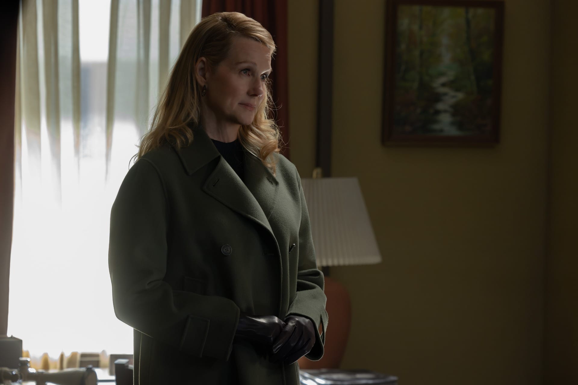 Production still of Lianna in a green coat standing in a room, a lamp and picture of a mountain stream behind her.