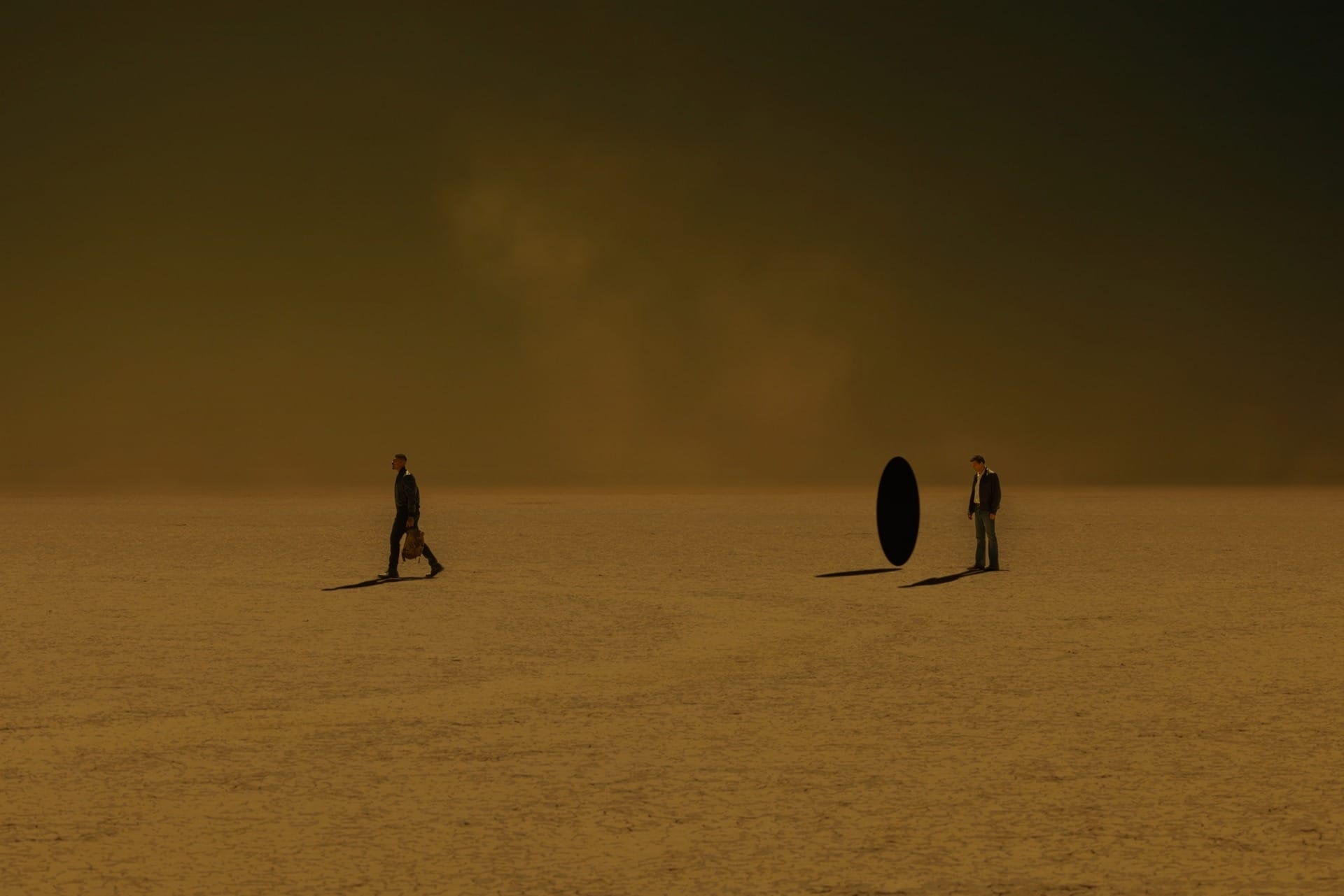 Production still of John and Hal on an alien planet. Hal stands beside a black portal. The landscape is aggressively brown and void-like.