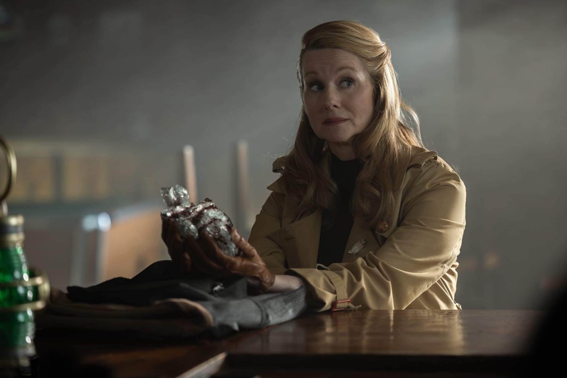 Production still of Lianna in a trenchcoat sitting at a bar and holding an item.