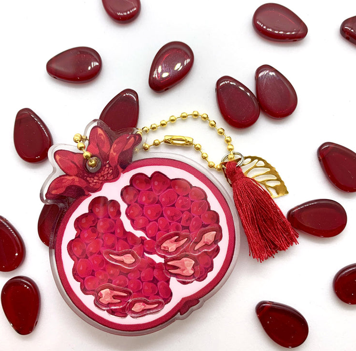 A red acrylic pomegranate charm with dynamic teeth inside that move when shaken. The background is white with small seedlike acrylic pips.