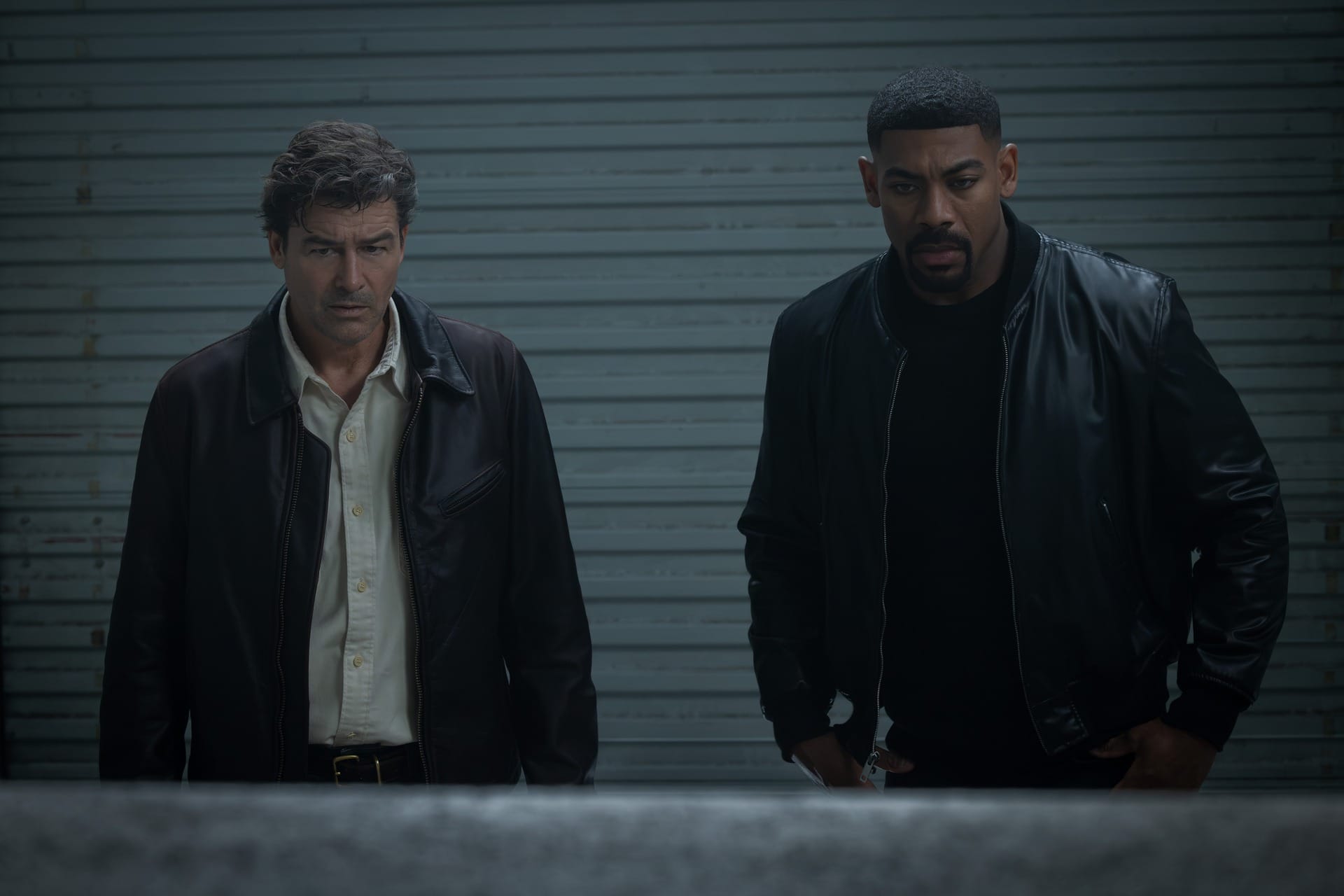 Production still featuring Hal and John backdropped by a greyish industrial wall/door.