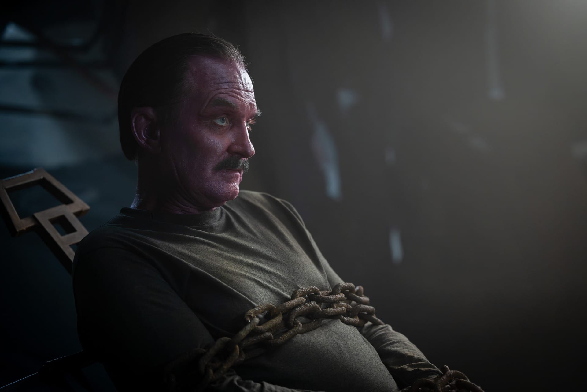 Production still of Sinestro (who does in fact look like an old white guy with a sunburn rather than a pink-skinned alien) dressed in greyish brown and chained to a chair.