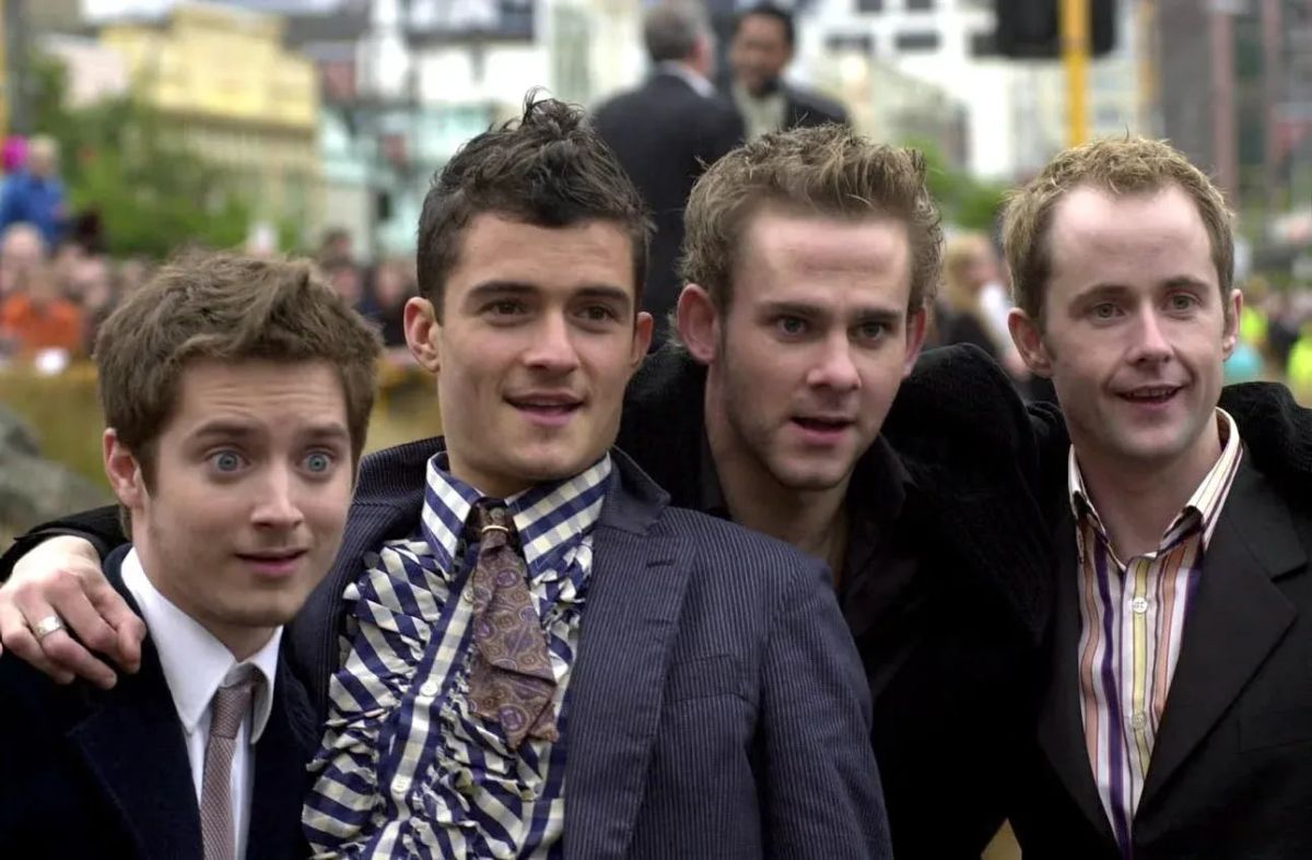 Photograph of actors Elijah Wood, Orlando Bloom, Dominic Monaghan, and Billy Boyd with their arms over each others' shoulders, posing for a photo.