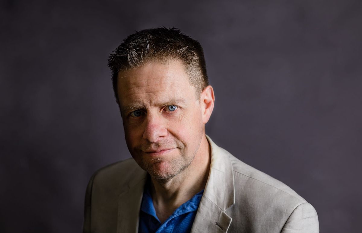 Headshot of Paul Cornell in a tan blazer and blue shirt, against a greyish brown background.