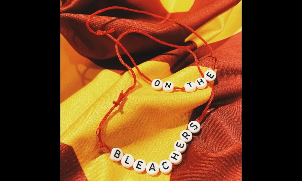 photograph of a red and yellow jersey with a beaded bracelet laid on top of it. Beads read: On the bleachers.