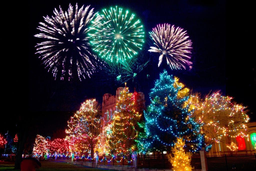 An image of fireworks over a set of buildings decorated in colorful Christmas lights.