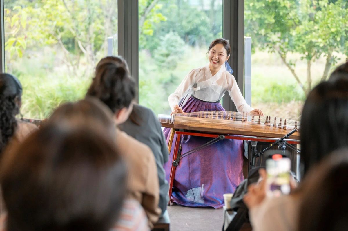 Photograph of Oh Hyun-ji playing gayageum in front of an audience, backdropped by green trees and bushes through floor to ceiling windows.