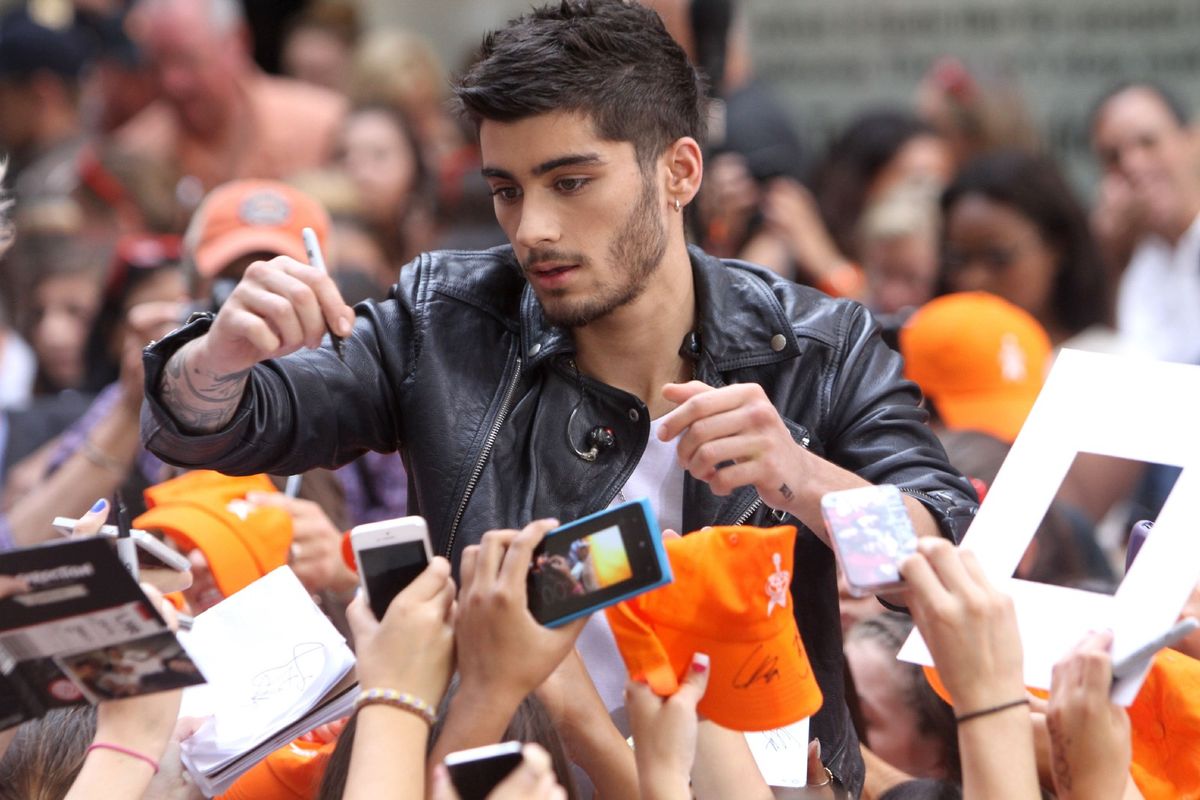 Photograph of Zayn in a leatherjacket standing in the center of a crowd of fans, holding a pen and poised to sign autographs. Fans are holding up phones, papers, and orange hats.